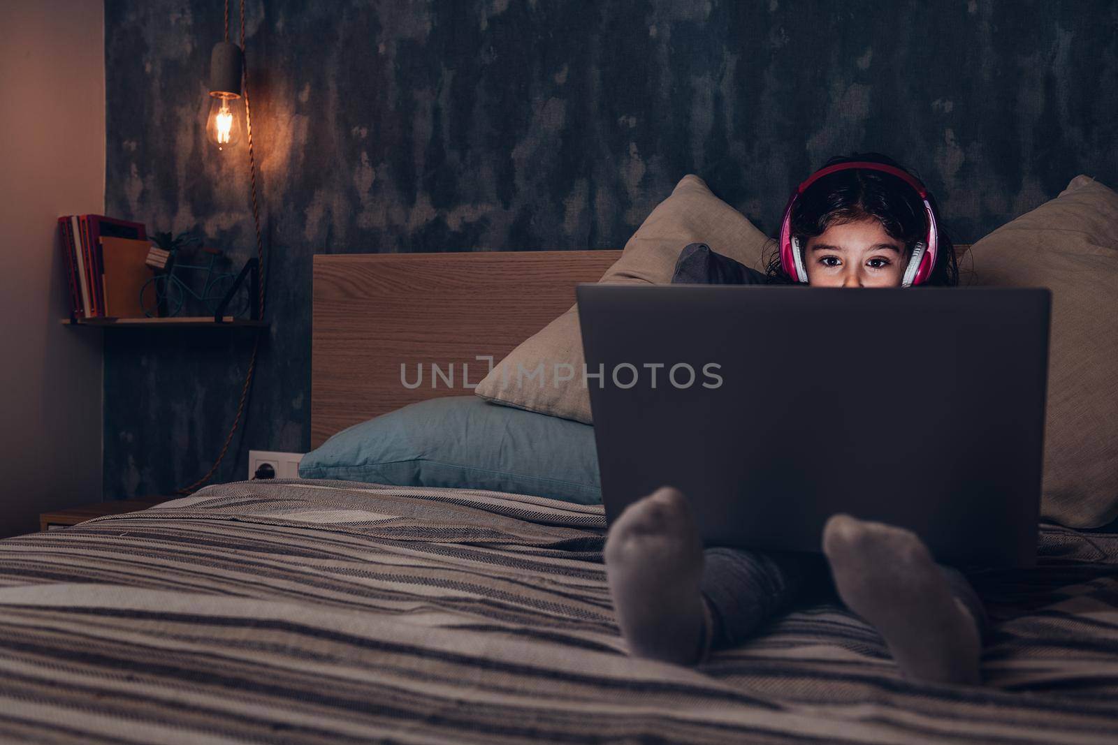 little girl sitting on the bed between cushions concentrated while playing or watching a movie on a laptop in the darkness, has a pink headset, child and technology concept, copy space for text
