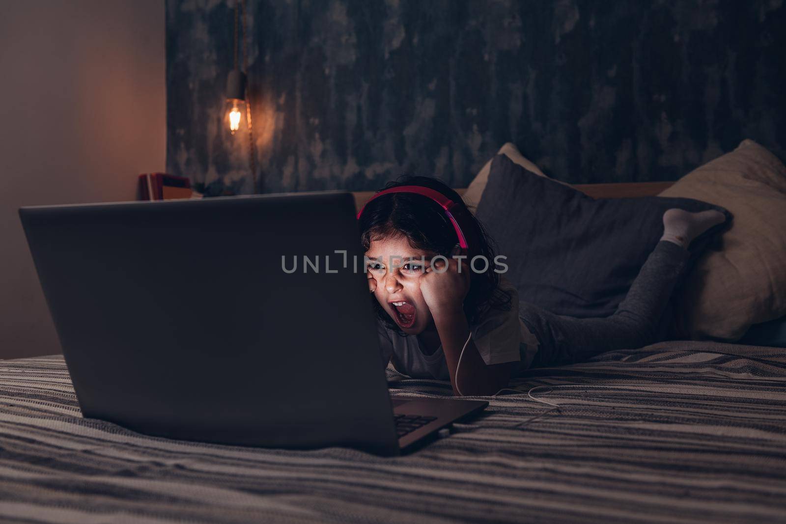little girl lying in bed screaming while watching a movie on a laptop in the darkness, has a pink headset, child and technology concept, copy space for text