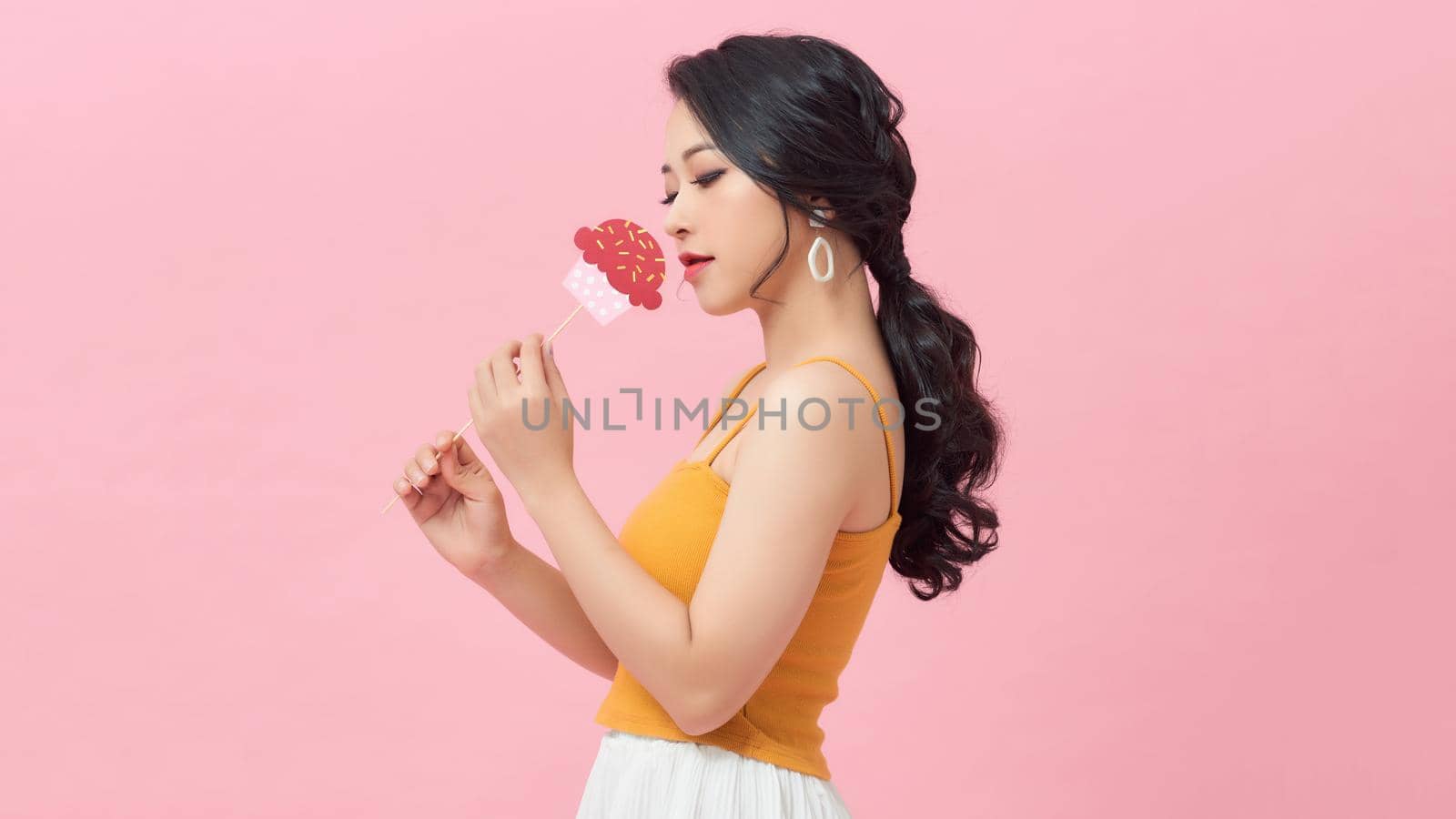 Happy young woman holding paper cupcake on stick over pink background