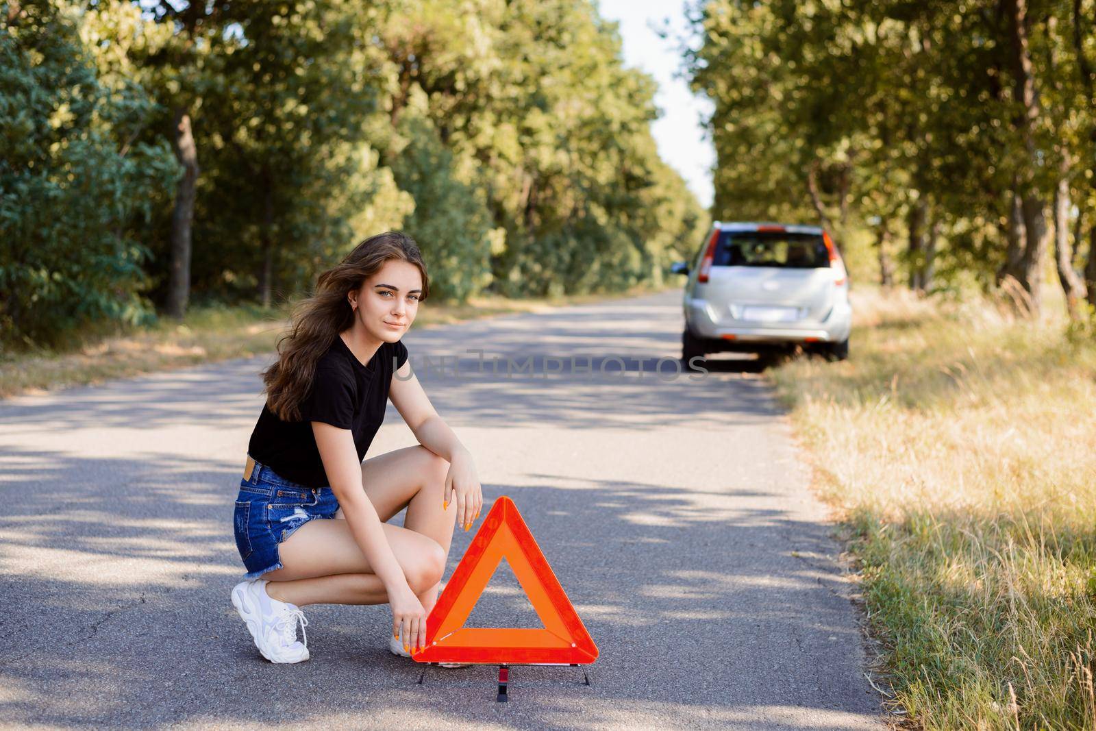 Sad young girl placing red emergency stop sign near broken silver car on the road