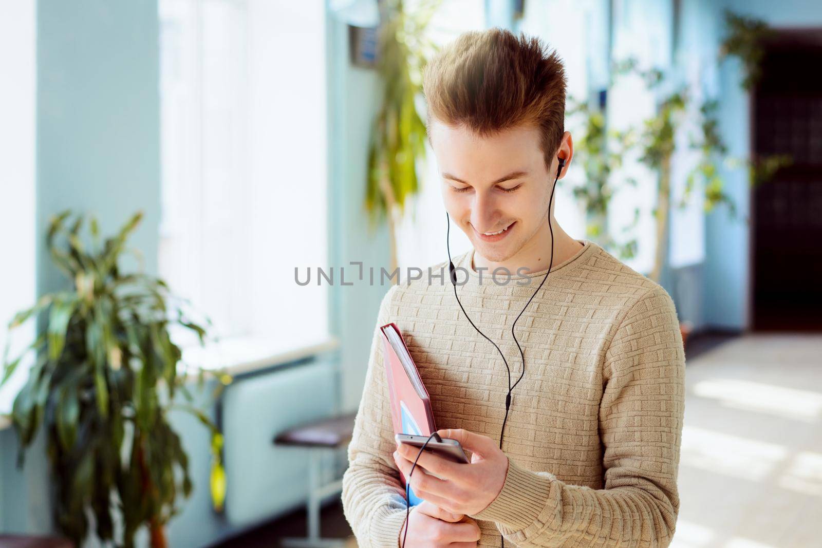 Happy male student using mobile phone and listens to music standing in campus with folder