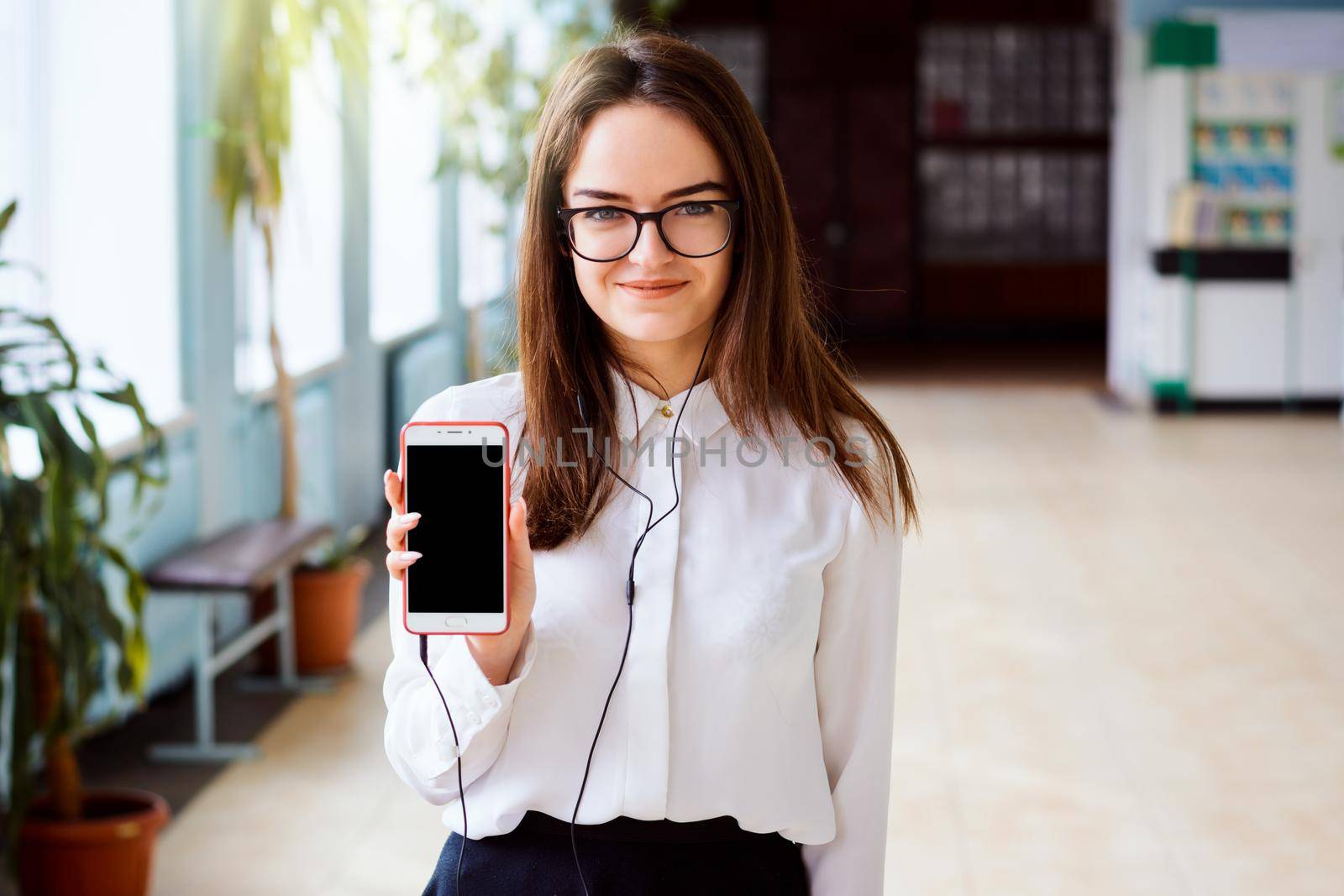 Portrait of a happy cheerful girl student with headphones holding black screen mobile phone and smiling to the camera over background of college corridor