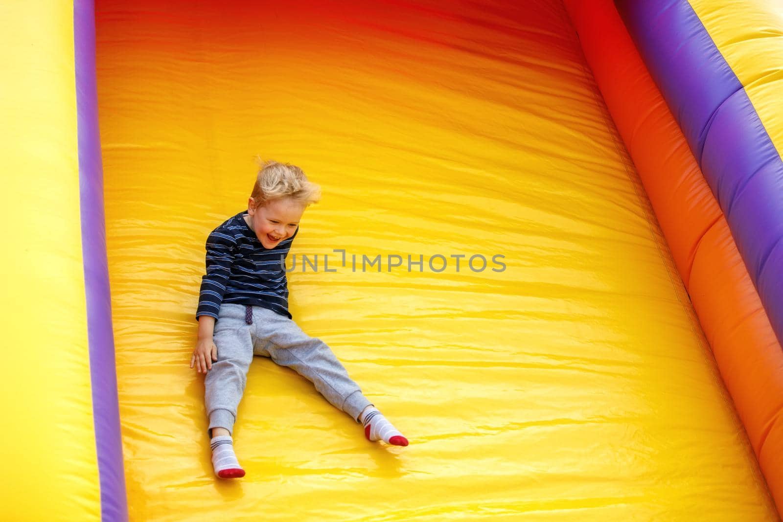 Laughing, lot of fun little boy child sliding on an inflatable multi-colored slide. Bright yellow background, free space for text by Lincikas