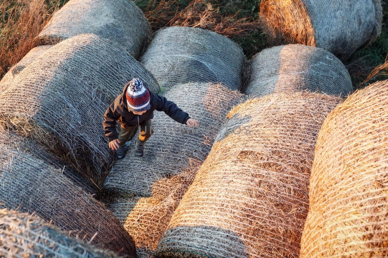 The little boy, in the autumn evening, in the sunset light, climbs up the hay rolls. Photographed from above by Lincikas