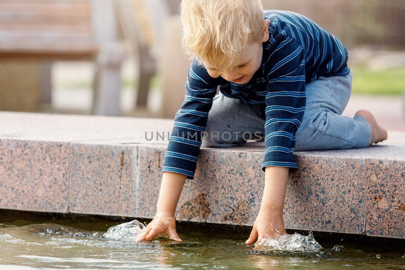 Happy boy touching water in fountain. Boy rejoice in summer in a beautiful fountain. Active summer leisure for kids in the city.