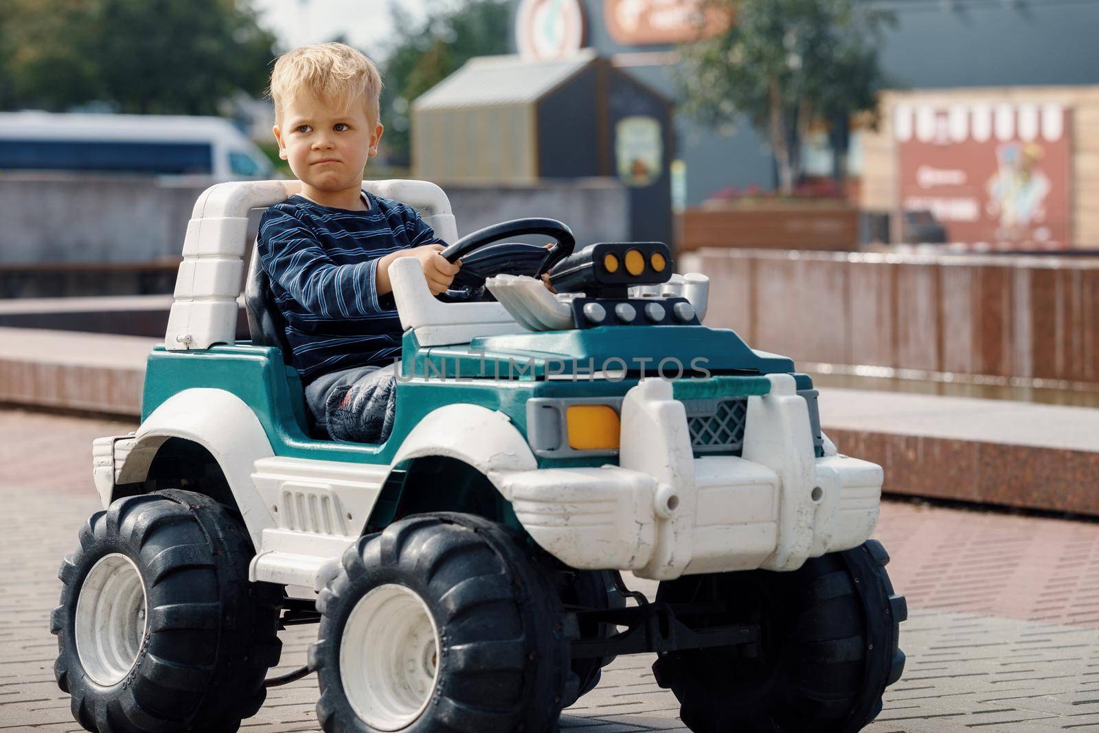 A relaxed, professional-faced cute little boy drives a large green toy electric SUV.
