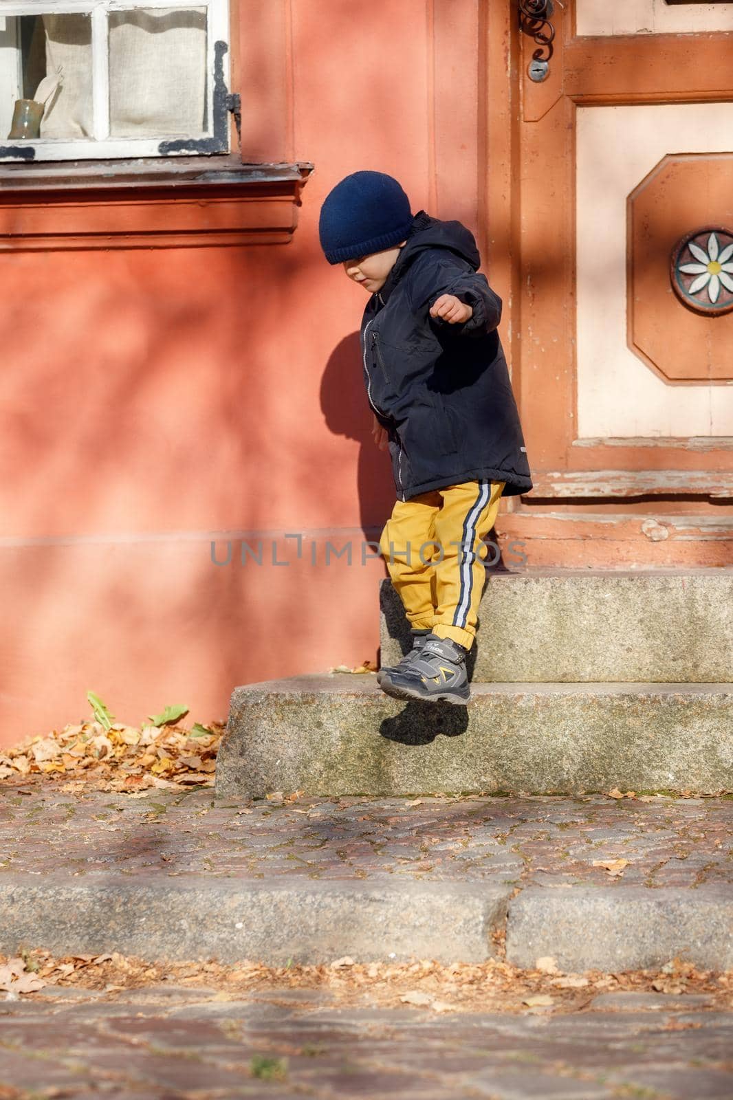 A cute boy in a autumn jacket with a blue hat goes for a walk around the old town. The child is self-climbing the stairs by Lincikas