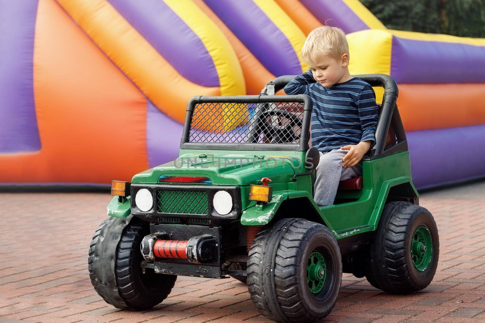 Little child driving green toy car, colored inflatable trampoline in background by Lincikas
