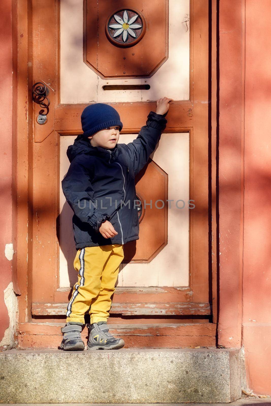 A cute little boy next to a big beautiful vintage door during a fall walk in the old town by Lincikas
