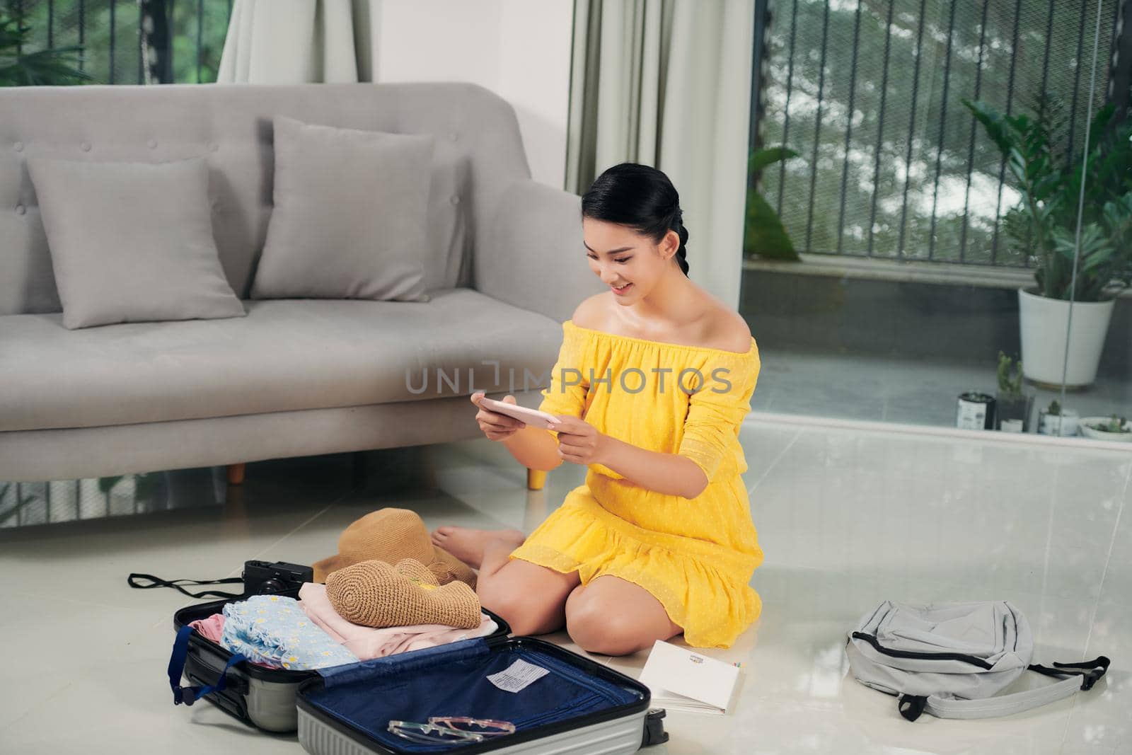 Young woman traveler using smartphone and packing her suitcase preparing for travel on summer vacation