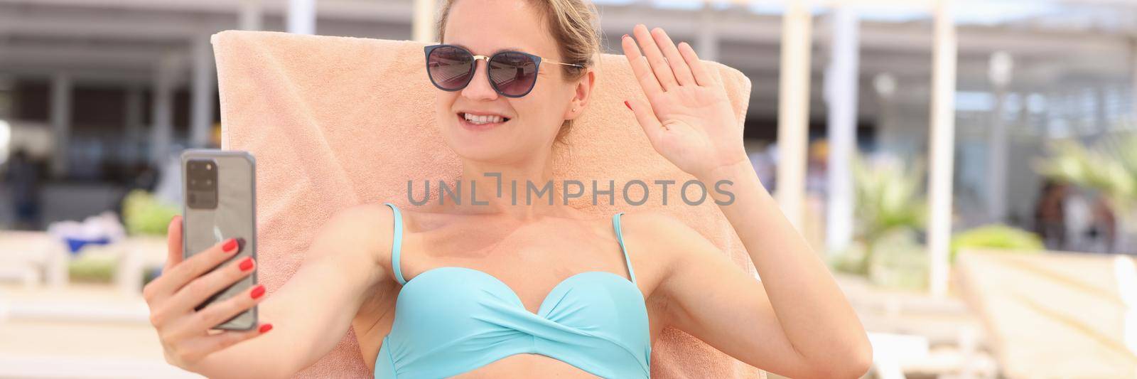 Woman in sunglasses lying on lounger and waving her hand at screen of mobile phone by kuprevich