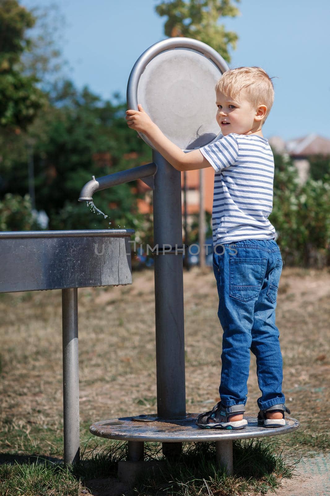 A happy little boy plays with a big water tap in a city park. Special water equipment for children's games on a hot summer day outdoors.