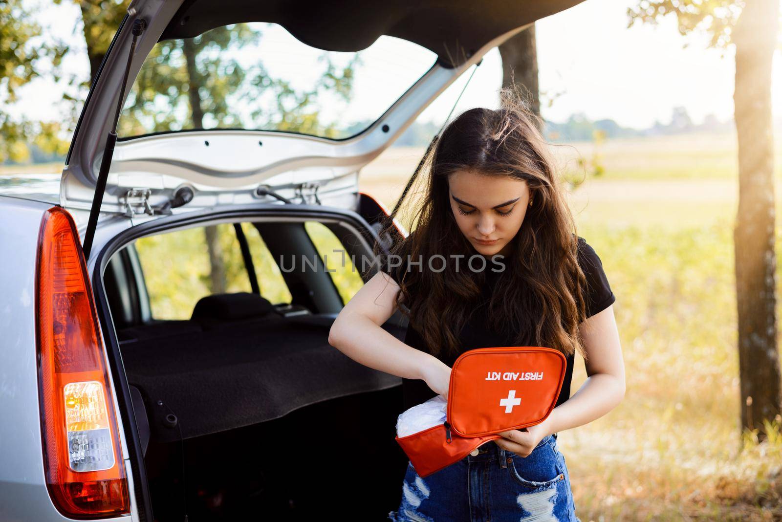 Young attractive girl stands near car with open back door and emergency light is on, tries to find something in first aid kit