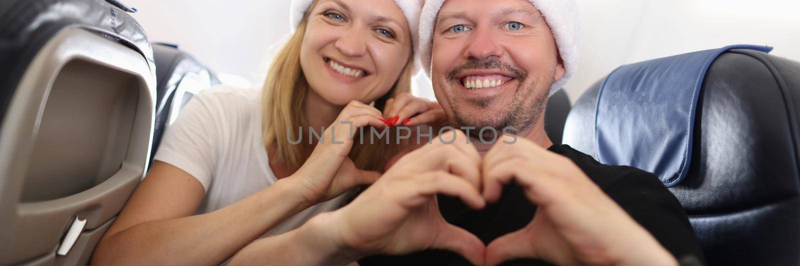 Man and woman in santa hats flying in airplane and showing heart with hands. Family new year holidays abroad concept