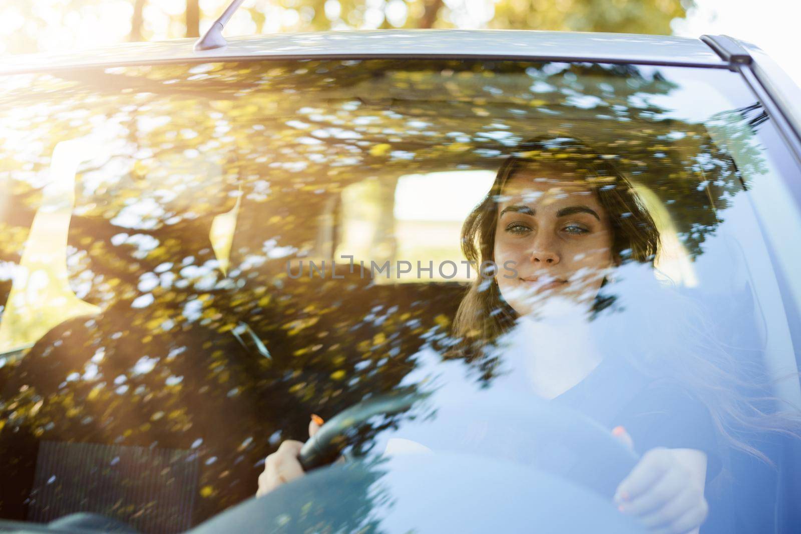Photo of a female student in driver`s seat while driving a car in countryside in the evening against setting sun