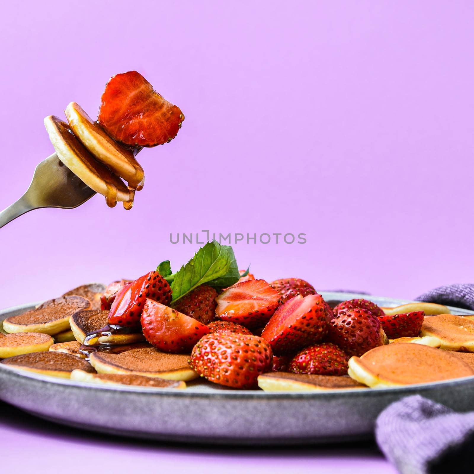 Selective focus on Maple syrup pouring on fork with tiny pancakes and strawberry. Pancake porridge, mini pancakes cereal in a bowl. Trendy food. Copy space for text