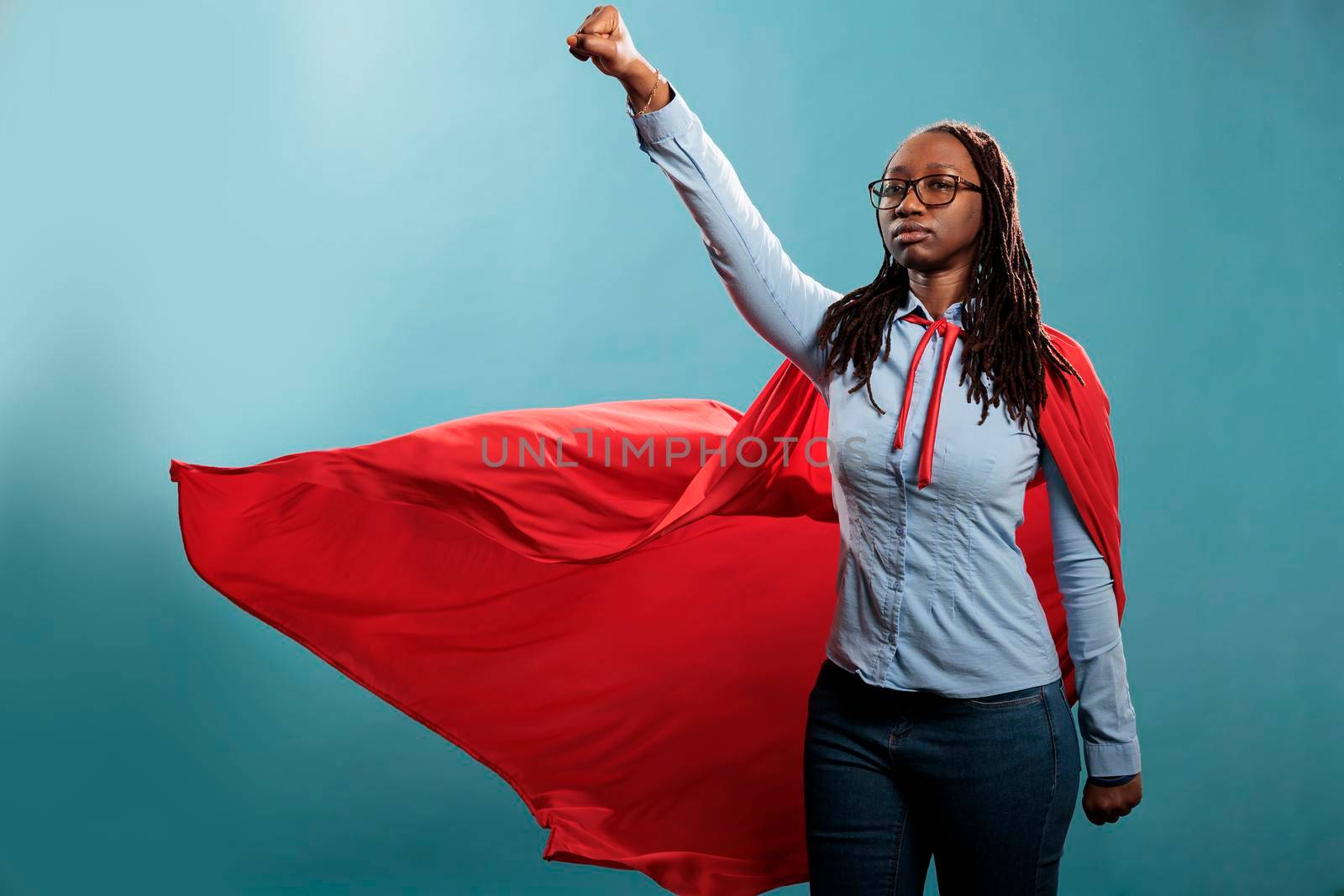 Powerful and brave young superhero woman wearing hero costume while posing as flying on blue background. Proud and strong african american justice defender looking ambitious while looking at camera.