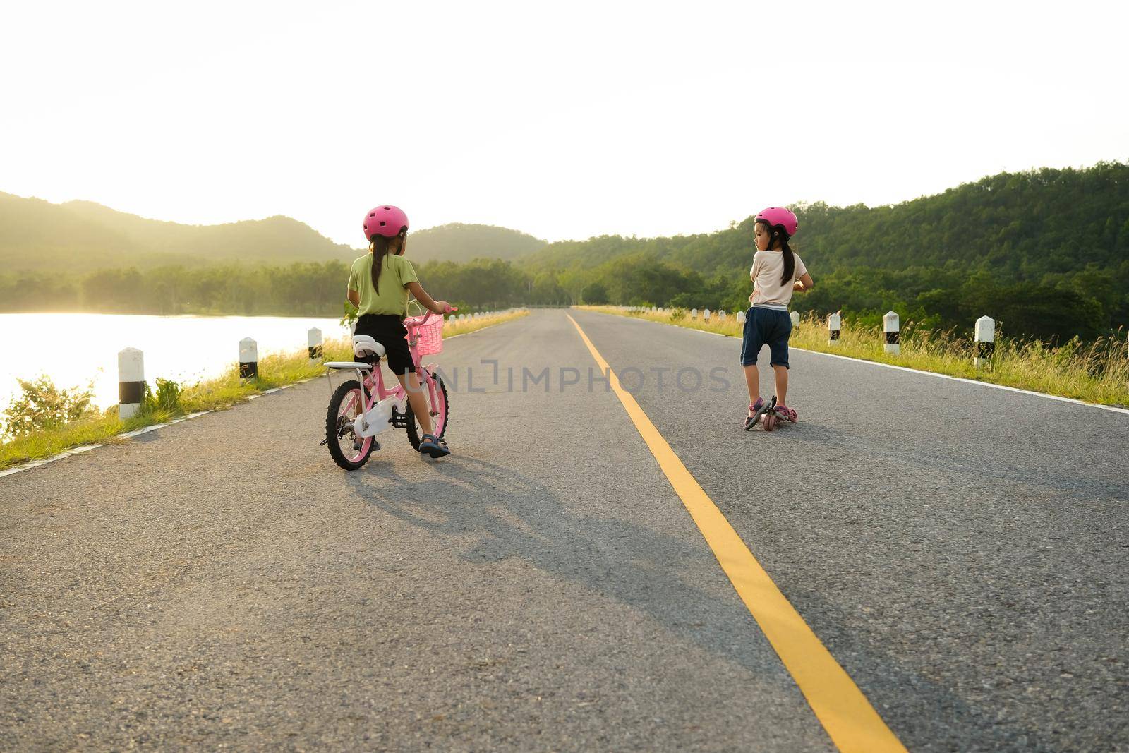 Cute little girl riding a bicycle and her sister riding a scooter on a lake road at sunset. Happy sisters doing outdoor activities together. Healthy Summer Activities for Kids. by TEERASAK