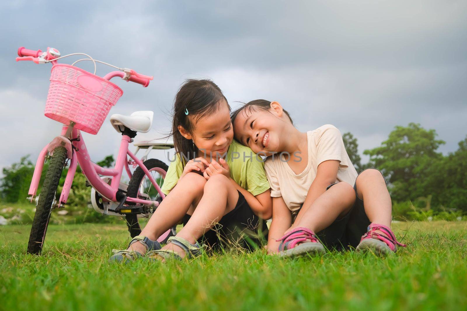 Happy cute little girl and her sister sitting on the lawn near the bikes in the park. Kids resting after biking. Healthy Summer Activities for Kids by TEERASAK