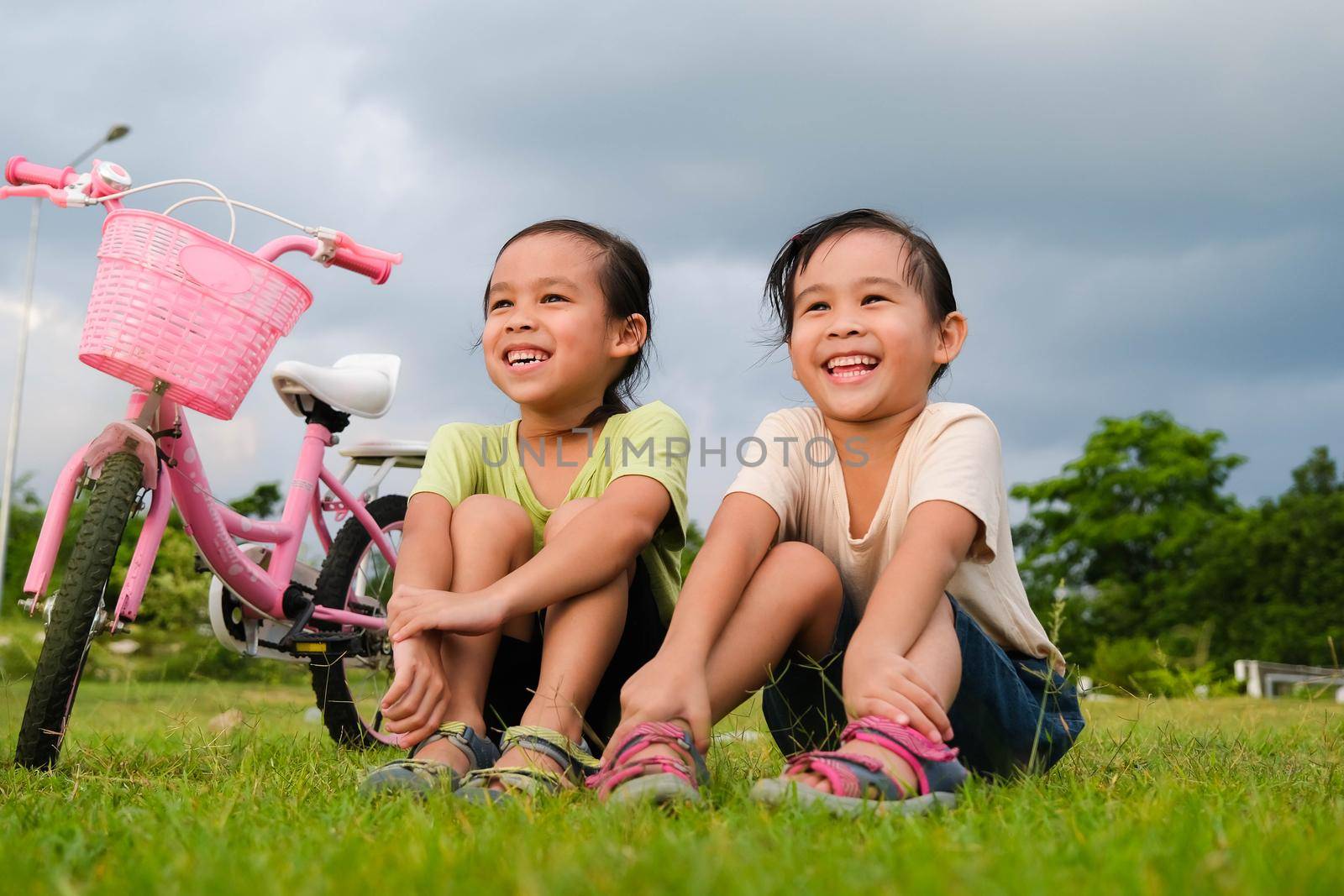 Happy cute little girl and her sister sitting on the lawn near the bikes in the park. Kids resting after biking. Healthy Summer Activities for Kids by TEERASAK