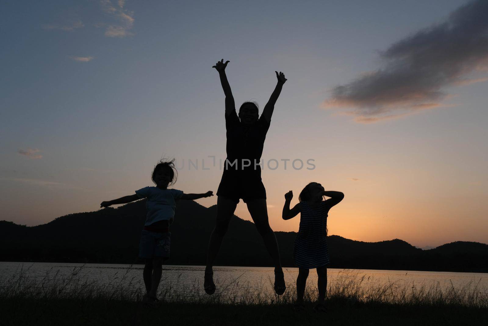 Silhouette of little daughters and mother jumping by the lake at sunset. Happy family playing together in the garden. Healthy summer activity for children. Funny time with family. by TEERASAK