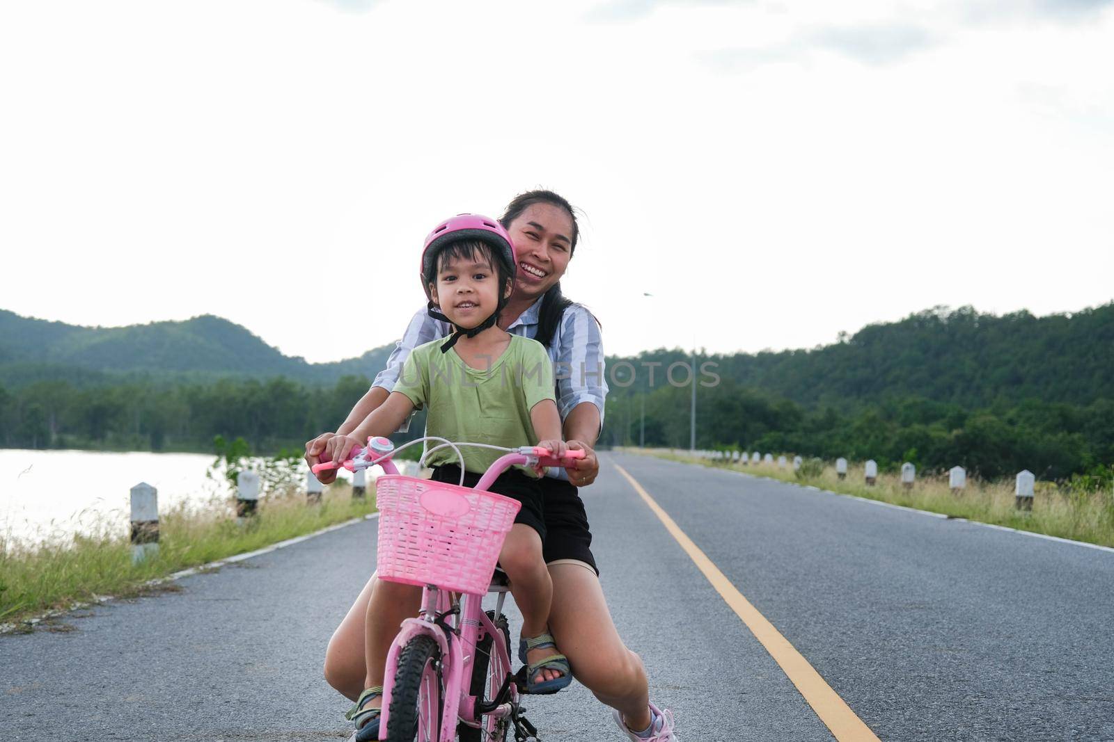 Cute little girl riding a bicycle with her mother on a lake road at sunset. Happy family doing outdoor activities together. Healthy Summer Activities for Kids.