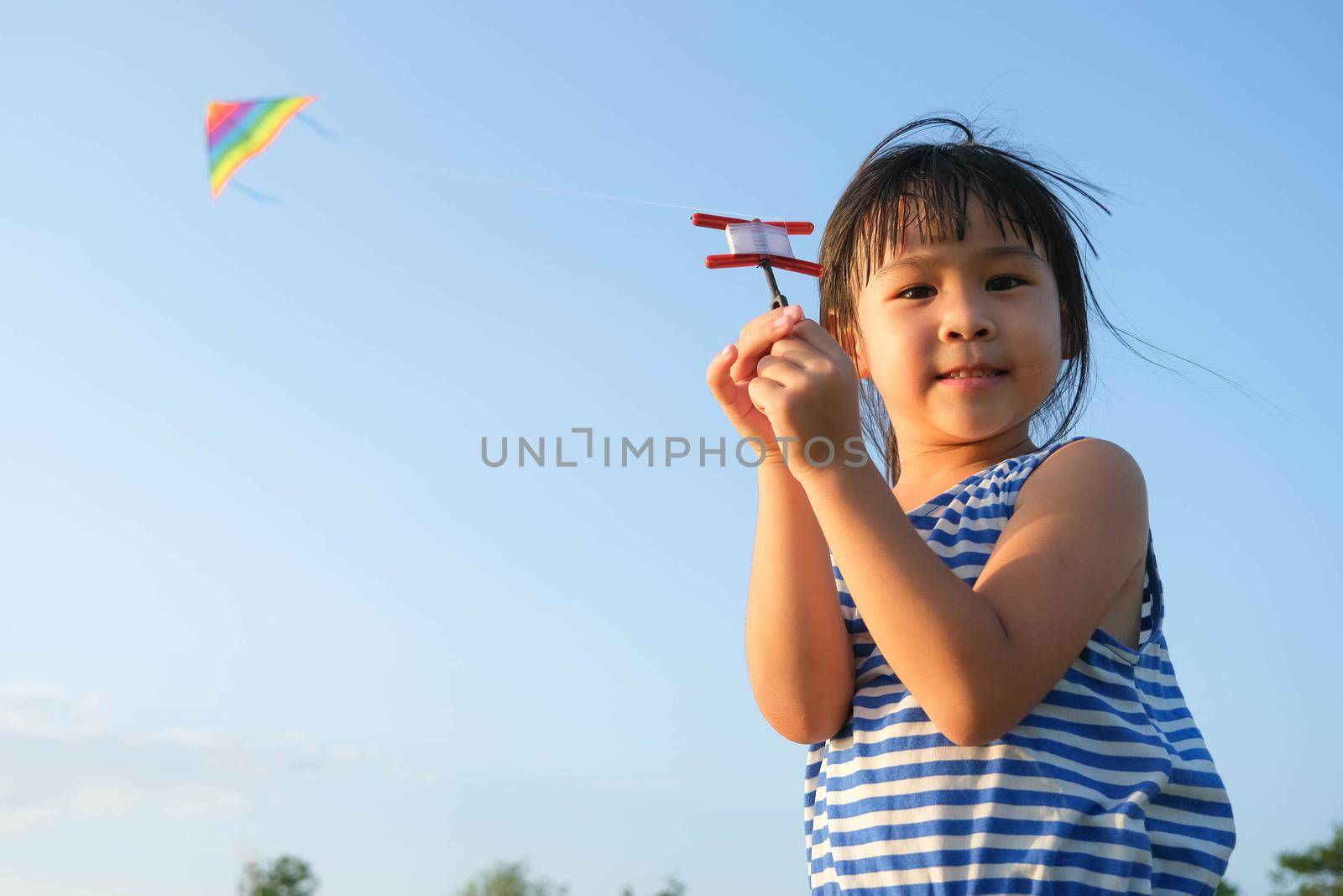 Child playing with a kite while running on a meadow by the lake at sunset. Healthy summer activity for children. Funny time with family. by TEERASAK