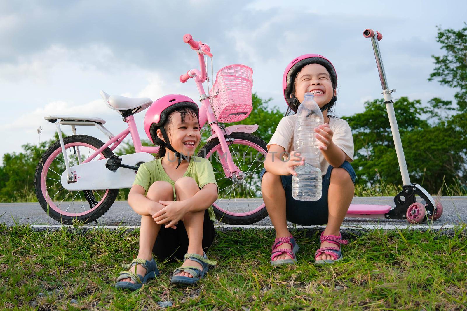 Happy cute little girl and her sister sit on the roadside lawn and drink water from a bottle near a bike in the park. Kids resting after biking. Healthy Summer Activities for Kids. by TEERASAK