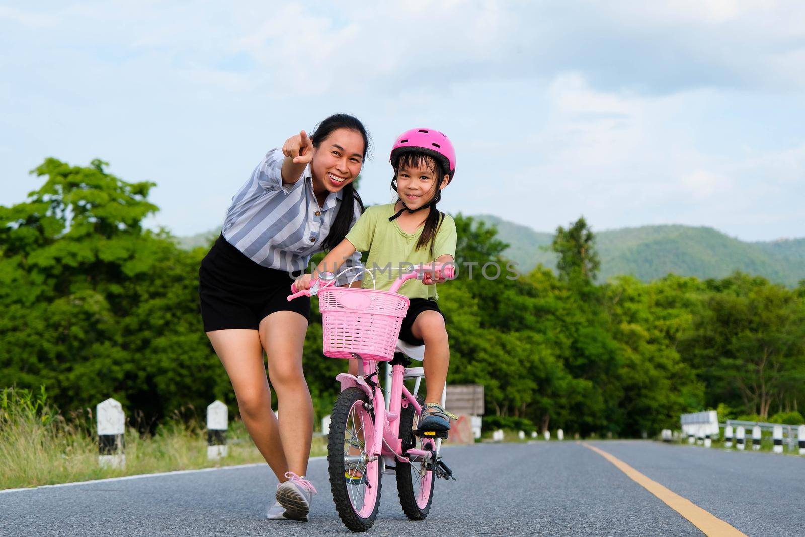 Asian mother teaching daughter to ride a bicycle in the park. A cute happy little girl learning to ride a bicycle with her mother. Active outdoor activities for kids. by TEERASAK