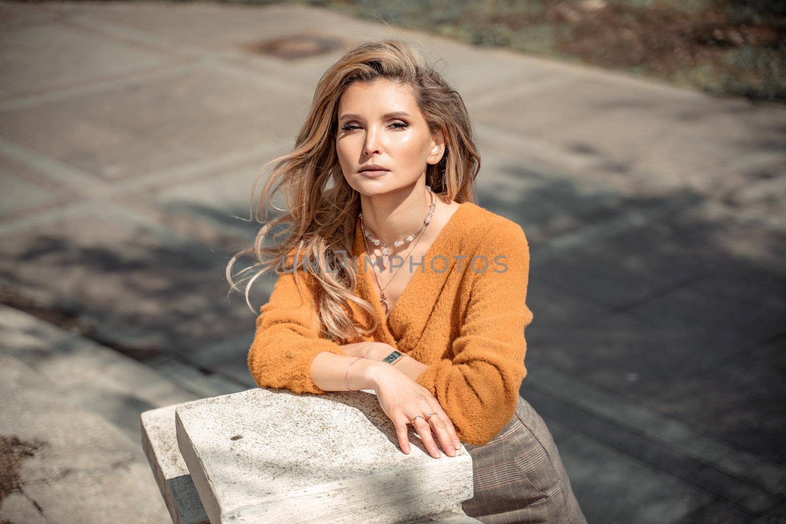 A middle-aged woman looks like a good blonde with curly beautiful hair and makeup on the background of the building. She is wearing a yellow sweater