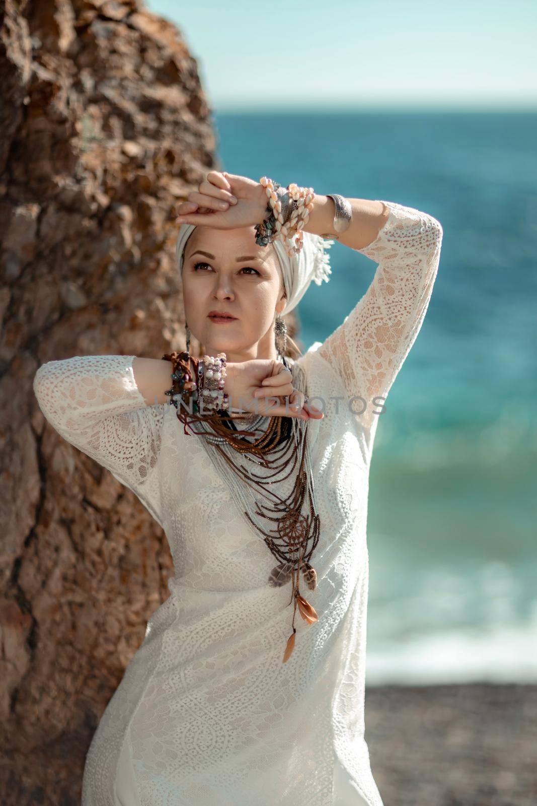 Middle aged woman looks good with blond hair, boho style in white long dress on the beach decorations on her neck and arms. by Matiunina