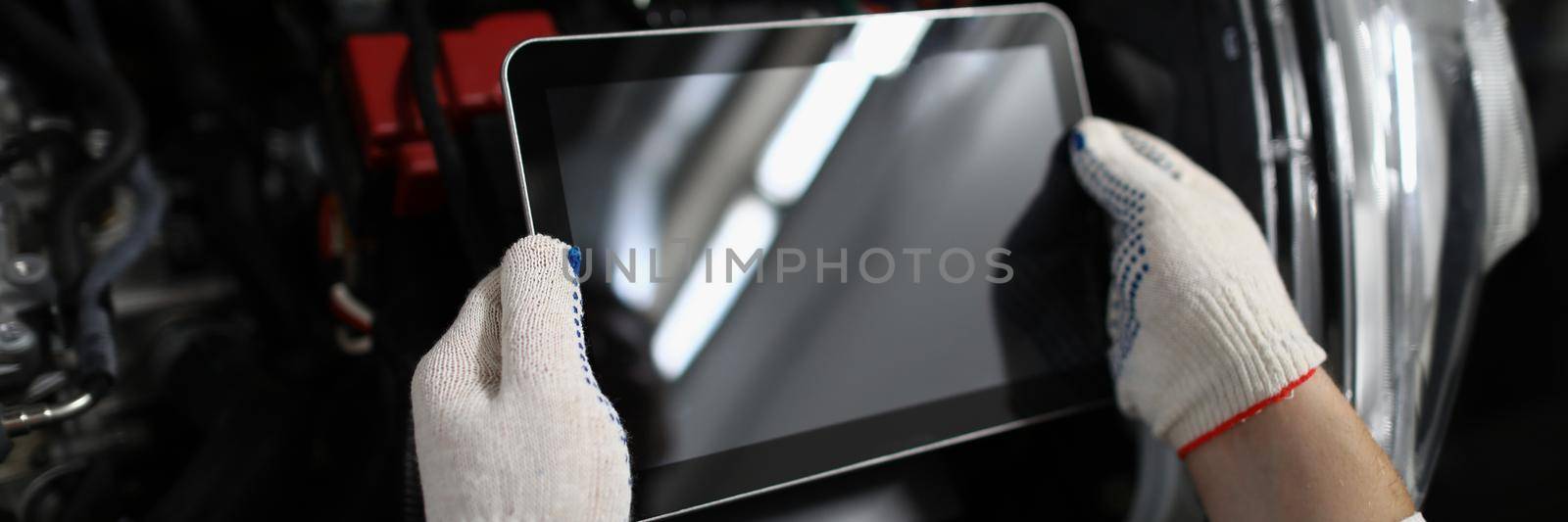 Man repairman holding digital tablet in front of open hood of car in garage closeup. Diagnostics of car breakdown concept