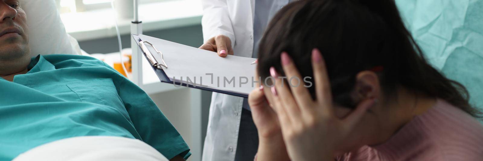 Doctor holding documents in front of crying wife and patient in hospital room. Support of incurable patients concept