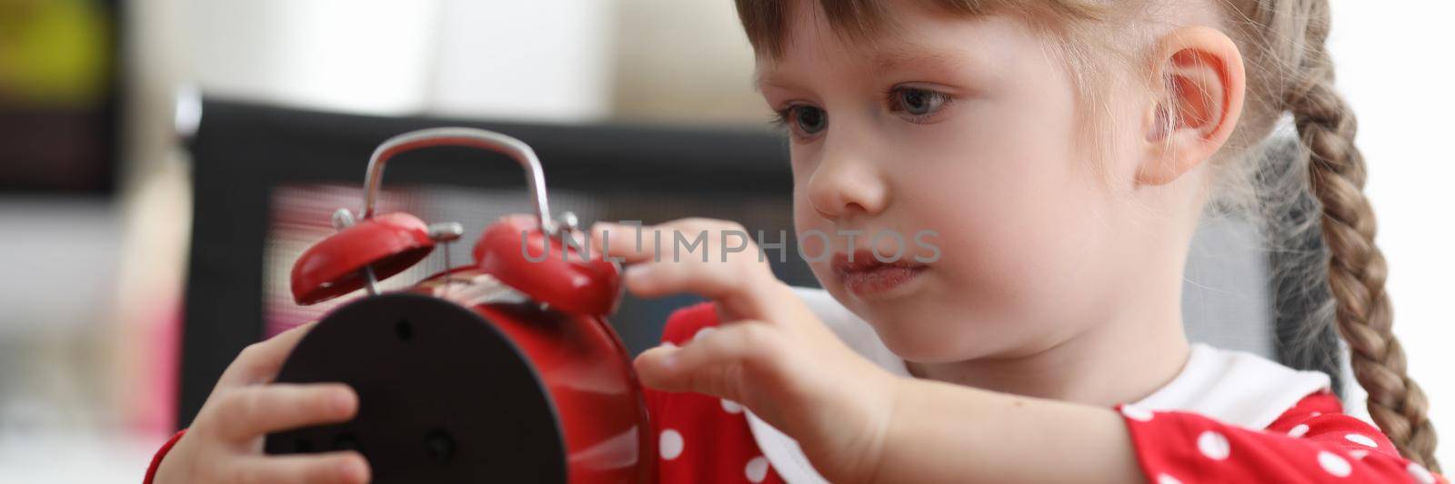 Little girl with pigtails looking at red alarm clock at table. Lesson schedule for kids concept