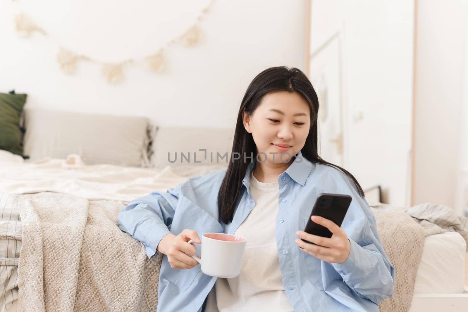 A portrait of a beautiful young Asian woman with a happy smile drinks morning coffee and looks at the mobile phone, sitting on the floor by the bed in the interior of the bedroom.