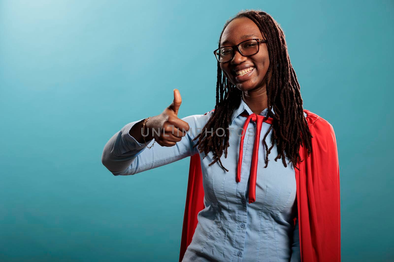 Young justice defender wearing red hero mighty cape showing thumbs up gesture sign at camera on blue background. by DCStudio