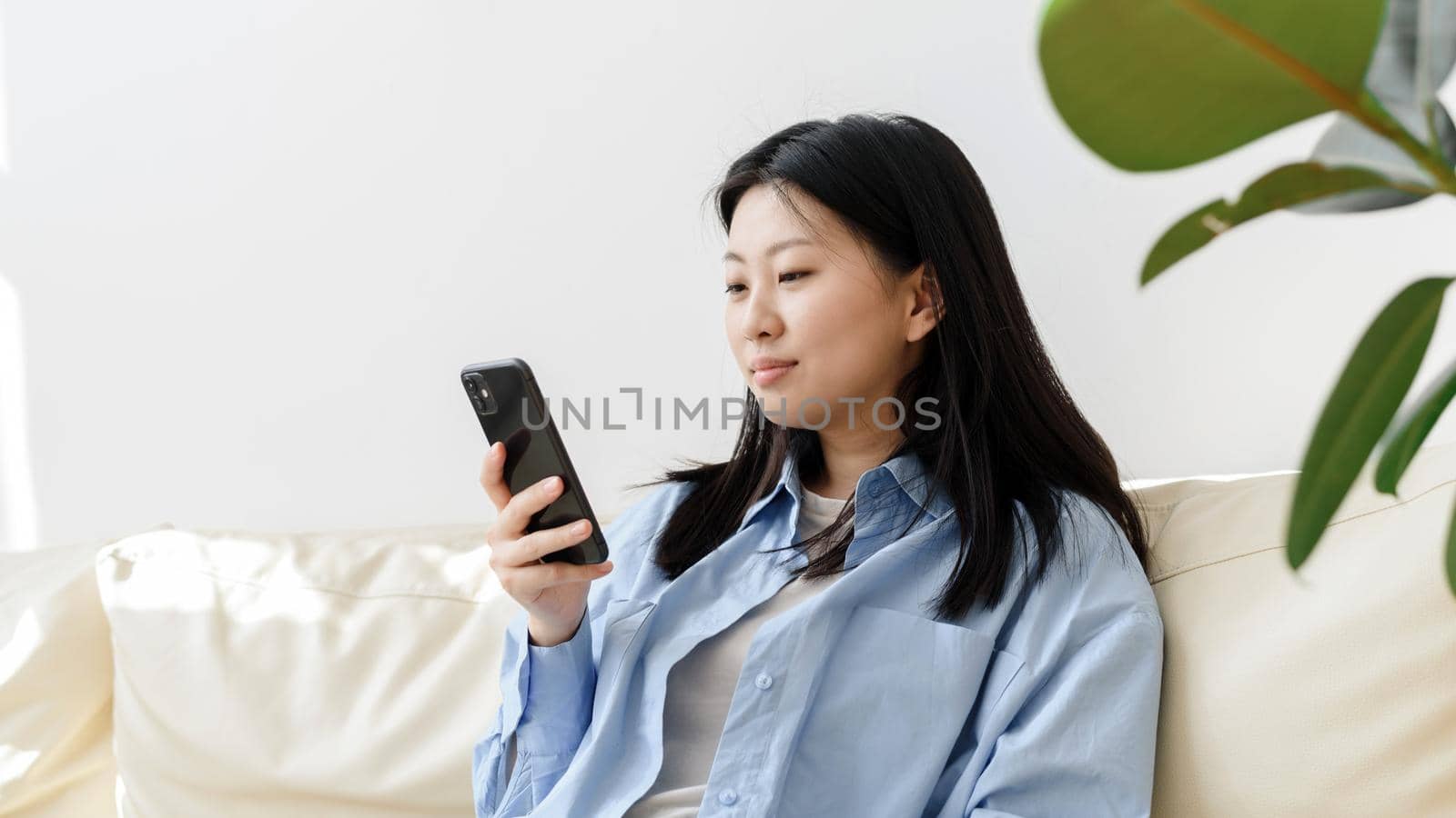 Young Asian woman relaxing on the sofa in the living room, browsing the news on her smartphone. Korean lady writes messages on modern mobile phone, shopping online through website. Web banner.