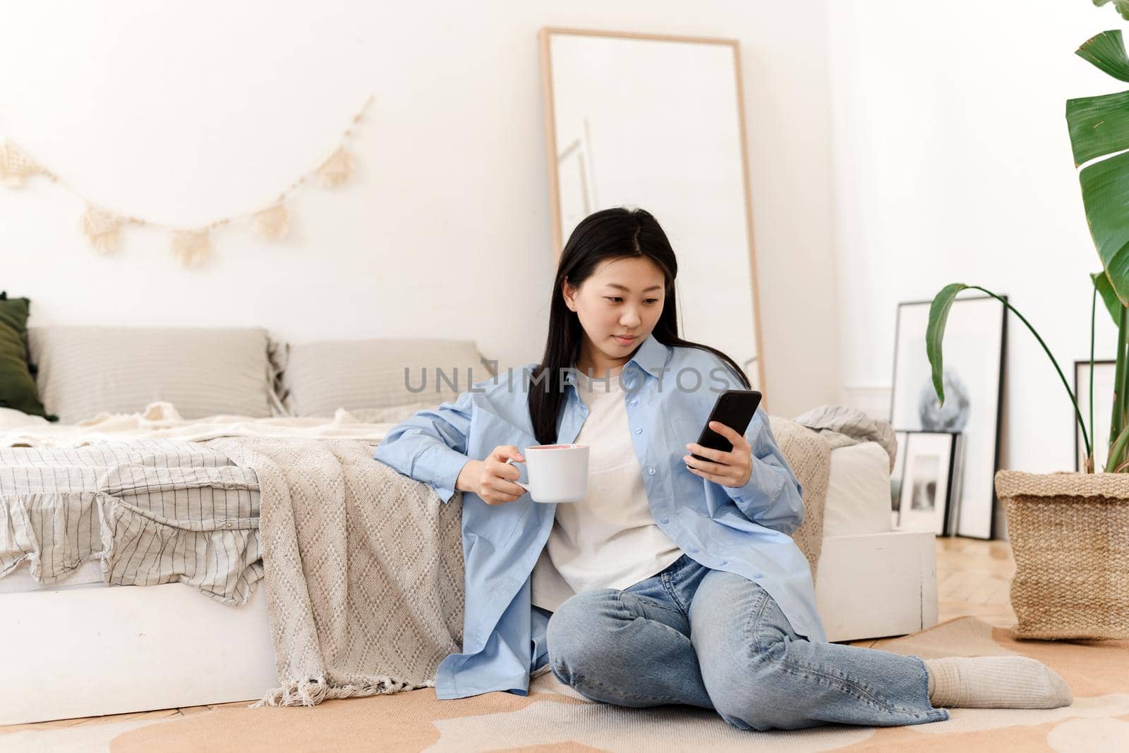 Photos of a charming young Asian woman sitting on the floor by the bed, drinking coffee and enjoying the weekend in the morning. A young Korean woman drinks warm tea, watching news on the phone