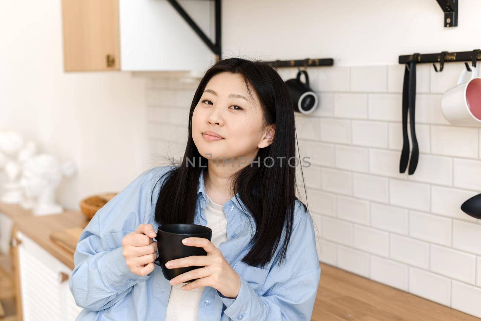 An attractive Asian woman rests in the kitchen, holds a mug with a hot drink and looks into the camera