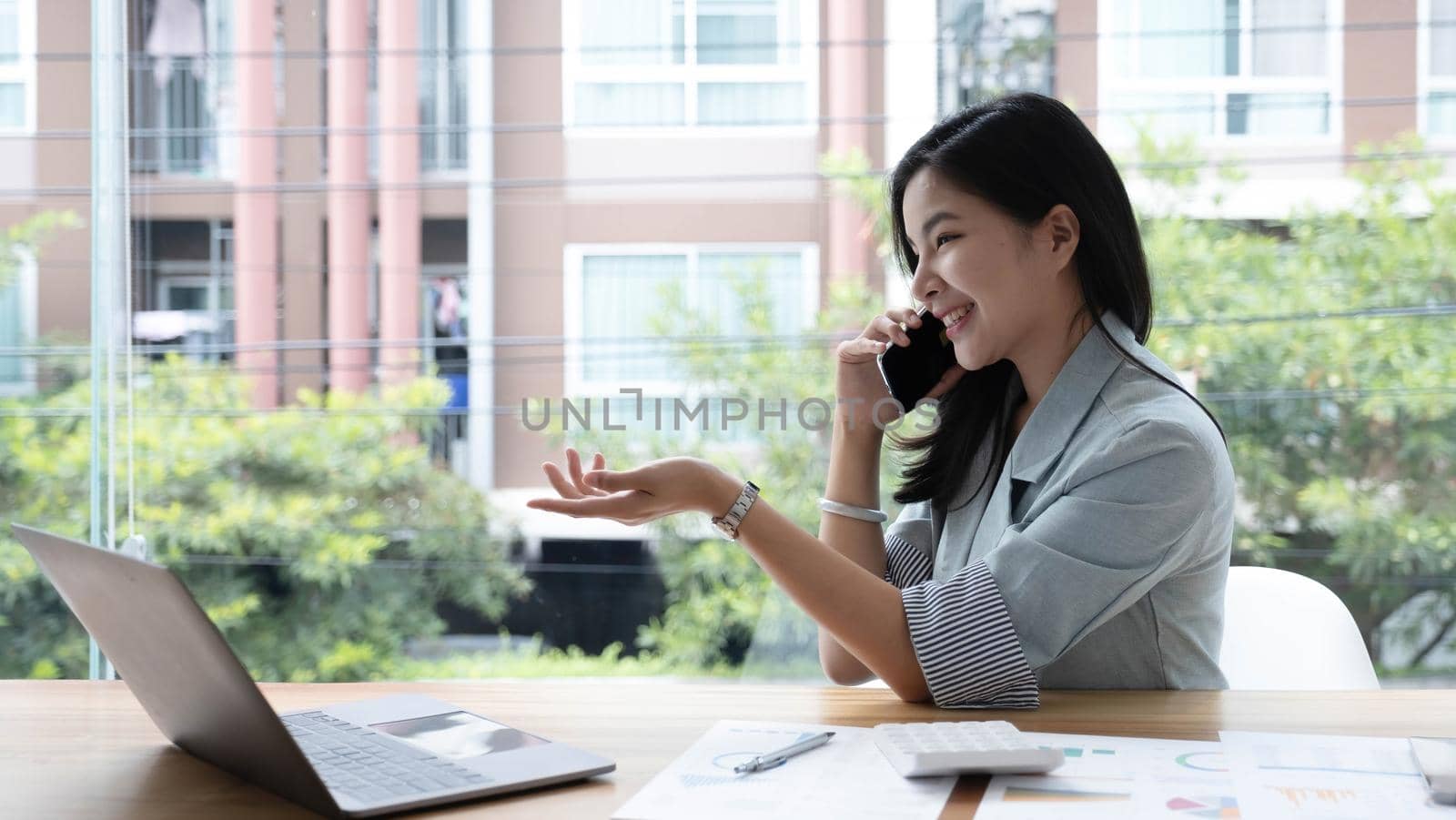 Attractive smiling young asian business woman relaxing at office, working on laptop computer, talking on mobile phone.
