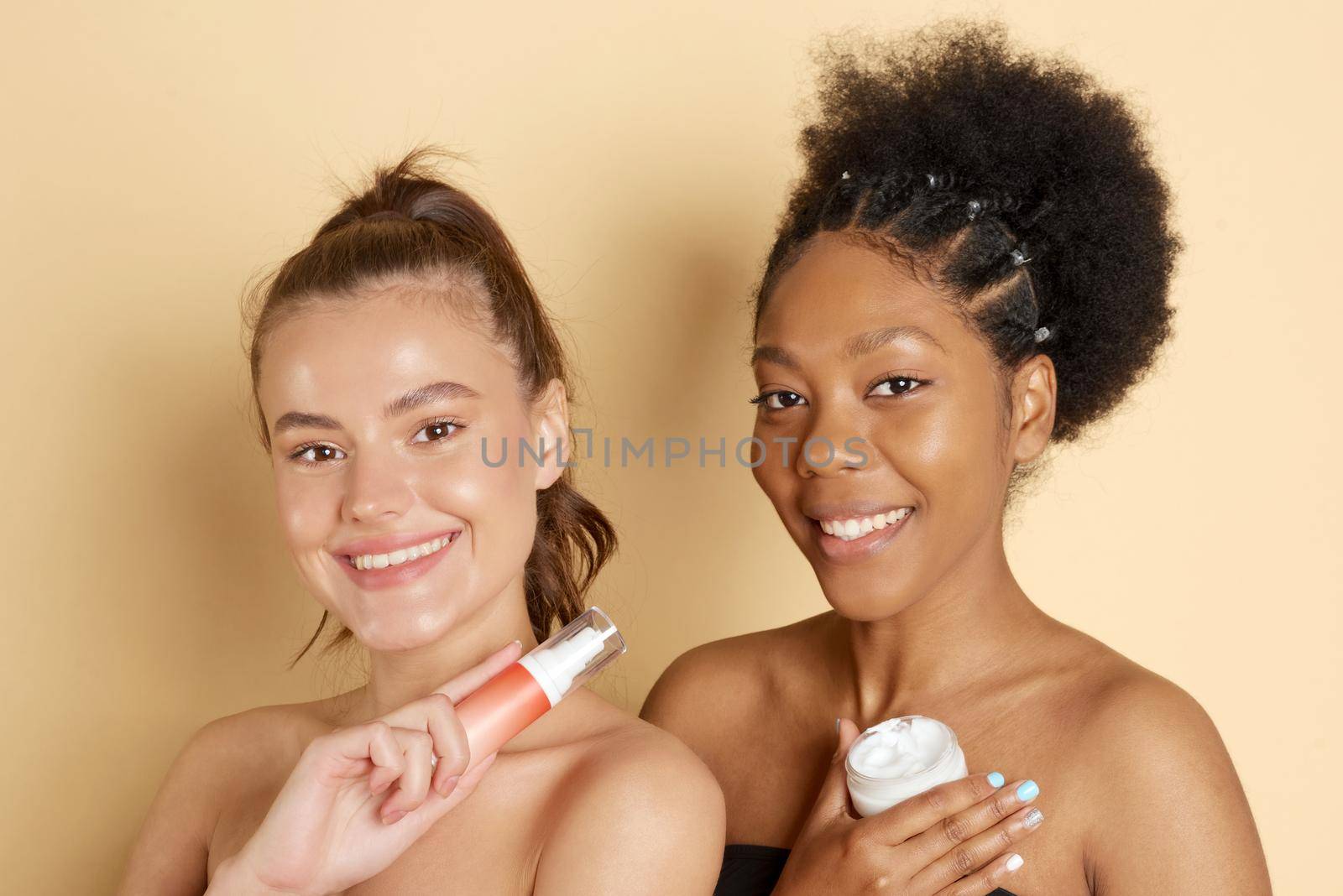 Smiling women hold jat of cosmetic moisturizer or face mask. Photo of attractive multiethnic women with perfect make-up on a beige background. Beauty concept.