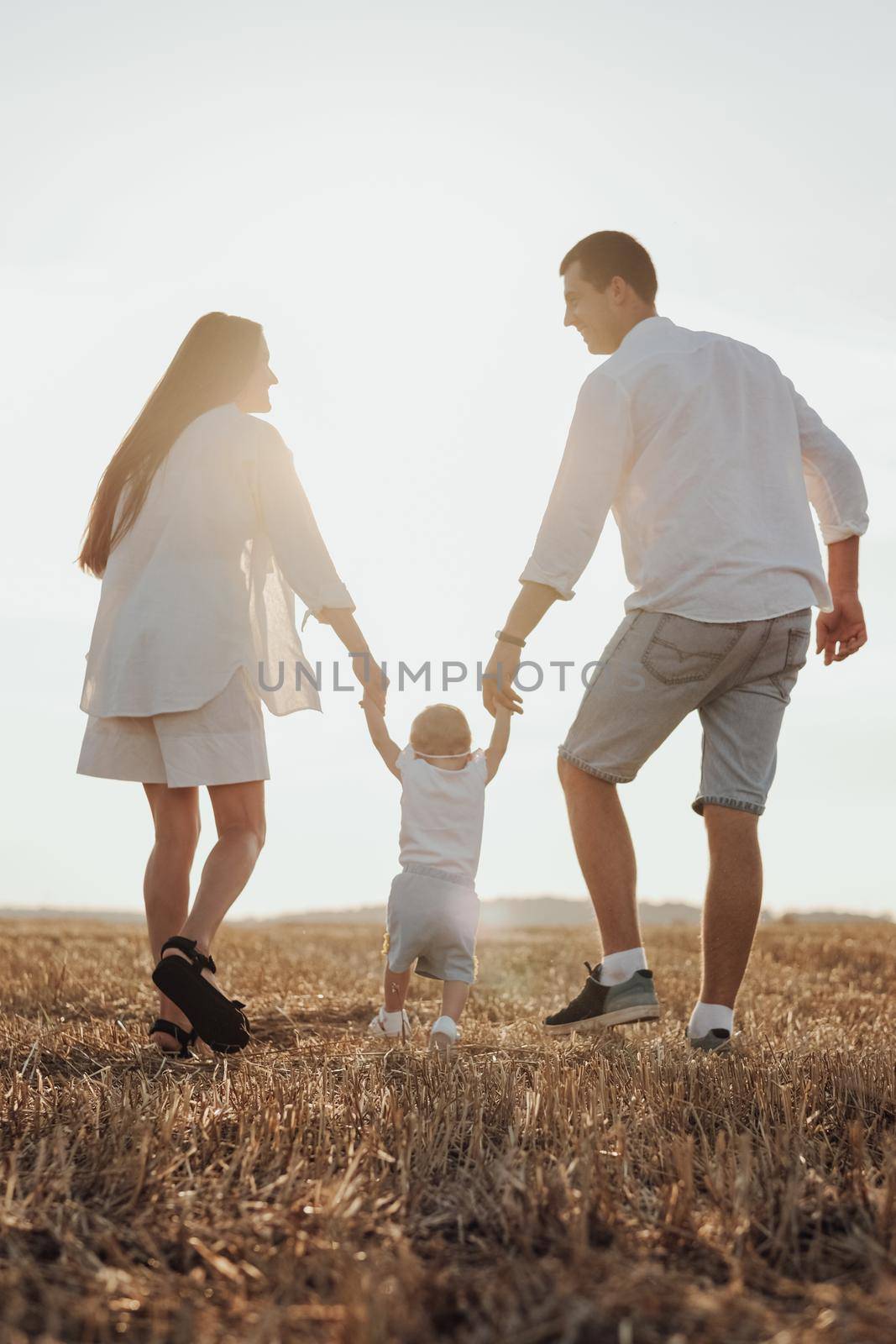Back View of Mom and Dad Helping Their Child Making First Steps, Young Family Walking on Field at Sunset