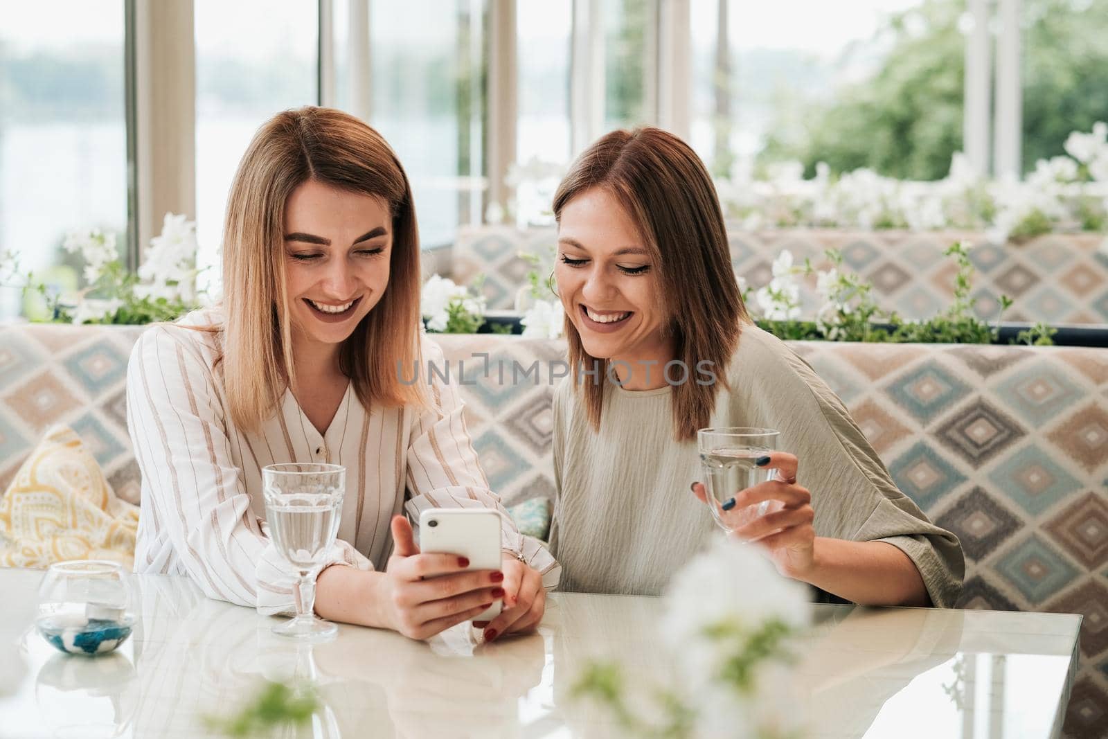 Two Cheerful Women Having Lunch Meeting in Restaurant, Watching on Smartphone and Smiling
