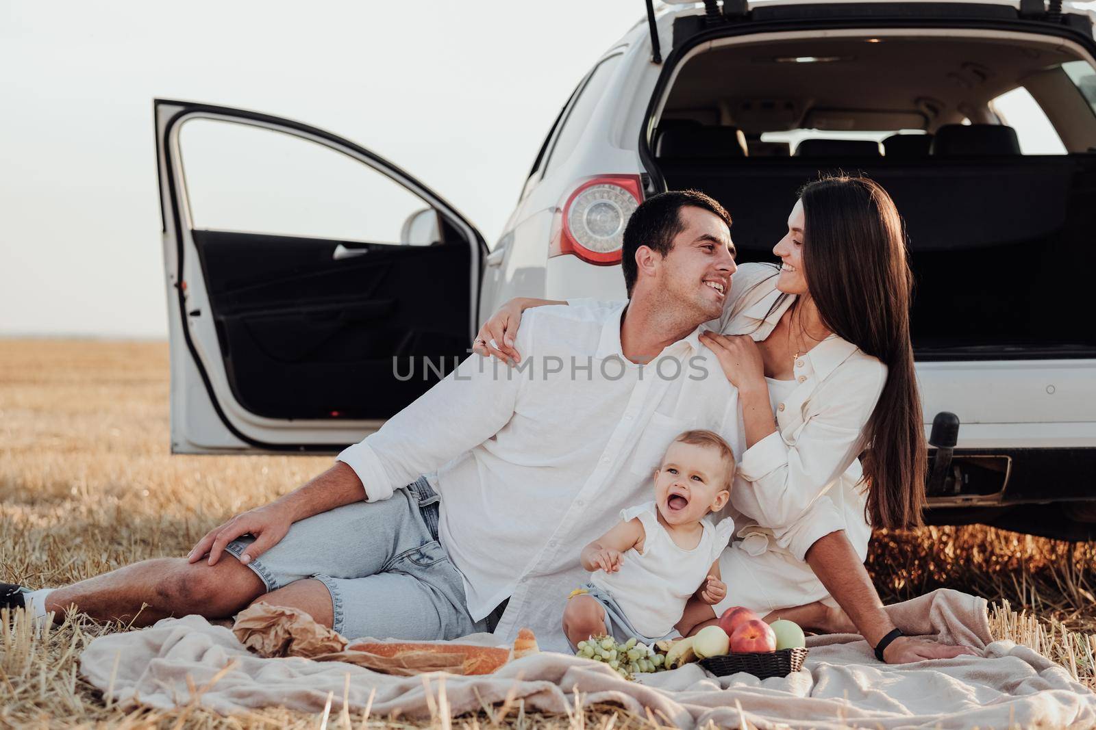Smiling Young Family with Toddler Child Having Picnic Outside the City, Mom and Dad with Their Daughter Dressed Alike Sitting Near Car Outdoors