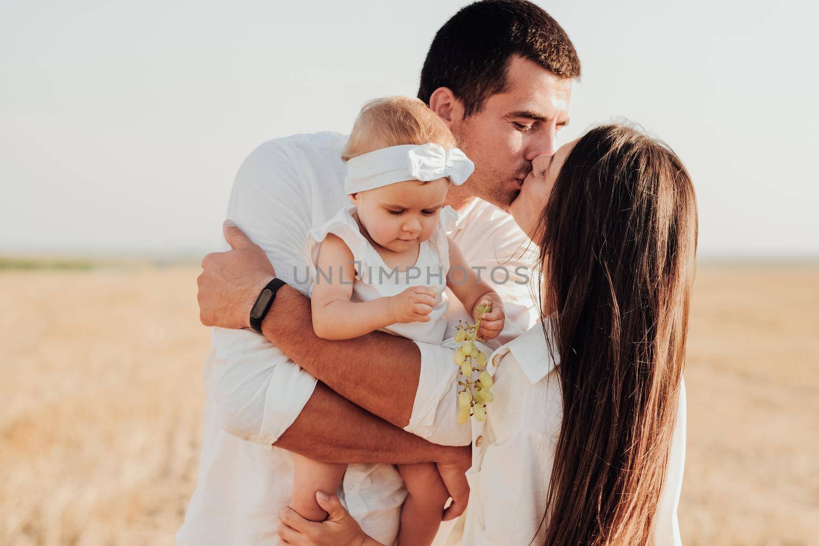 Young Caucasian Family with Toddler Child Hugging Outdoors in Field, Mother and Father Kissing While Their Daughter Eating Grape