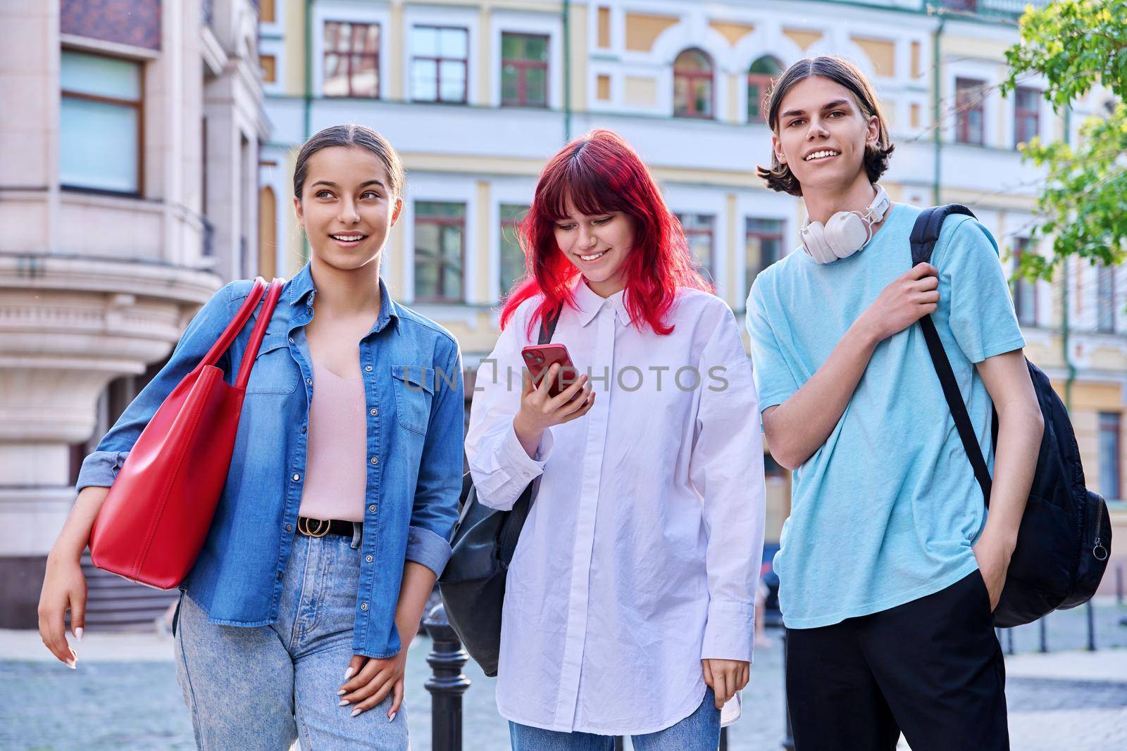 Three teenage friends met talking having fun outdoors. Smiling laughing teenagers with smartphone, together on city street. Communication, friendship, youth, lifestyle concept