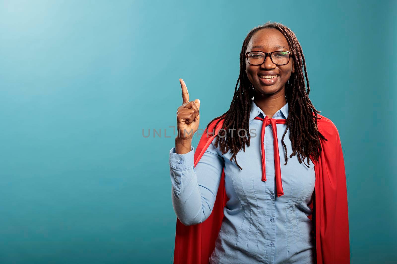 Optimistic happy superhero woman wearing red cape pointing finger up while standing on blue background. Proud and positive justice defender smiling at camera while gesturing fingers up. Studio shot