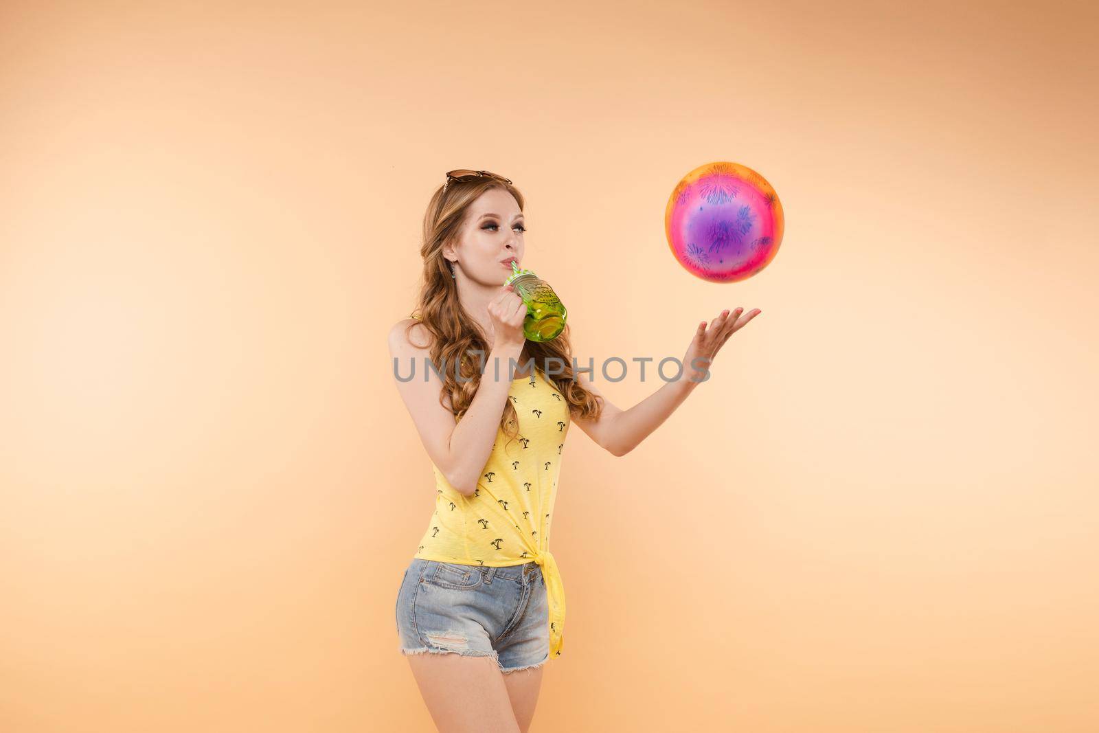 A woman in bright clothes, a yellow T-shirt and denim shorts, sunglasses holds a ball and cold lemonade in her hands, on an isolated background.