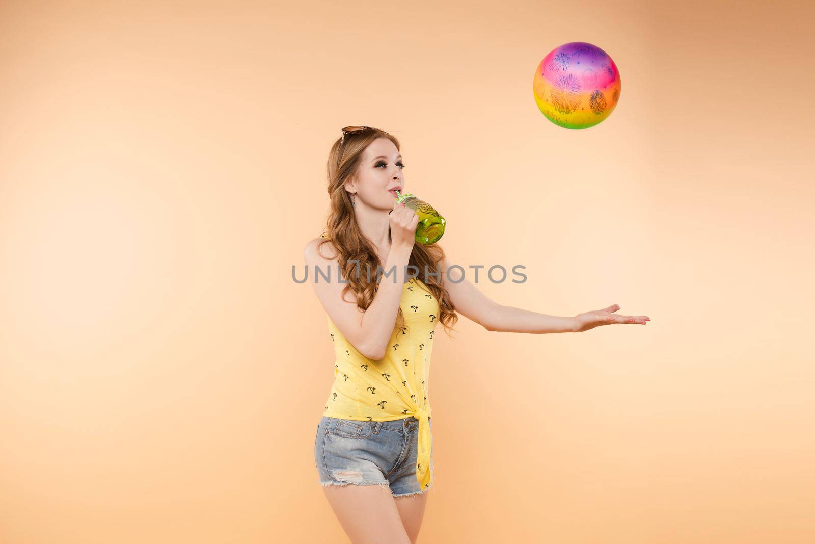 A woman in bright clothes, a yellow T-shirt and denim shorts, sunglasses holds a ball and cold lemonade in her hands, on an isolated background.