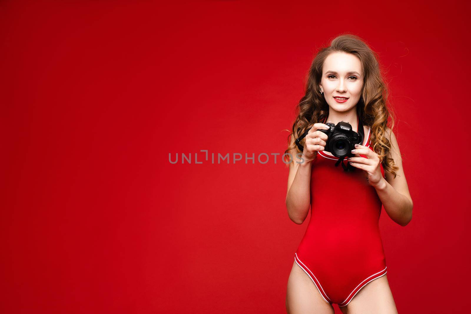 A beautiful woman in a red swimsuit with bright makeup and red lips with a camera in her hands.